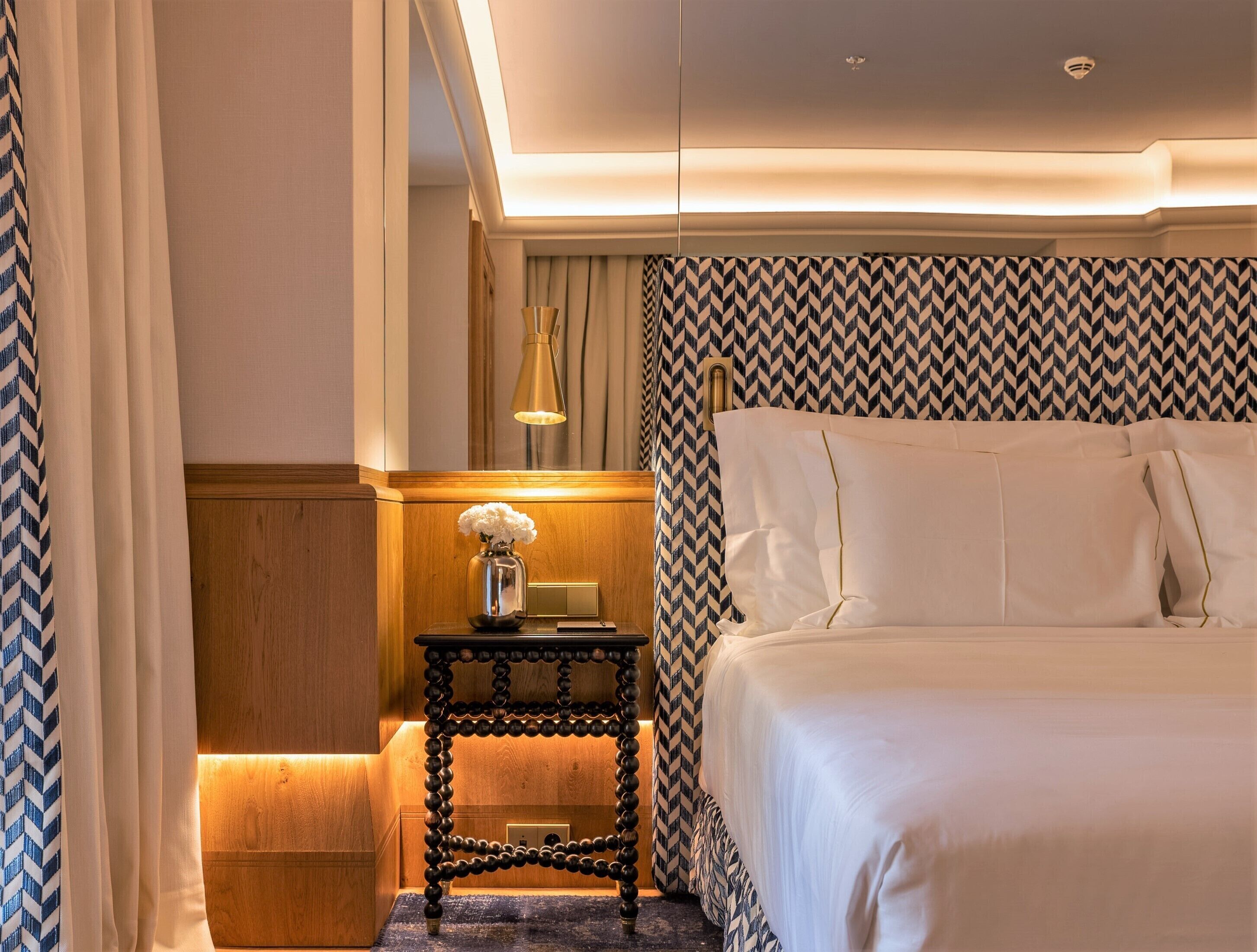 A hotel room with a bed, a nightstand with a lamp, and a patterned wall.