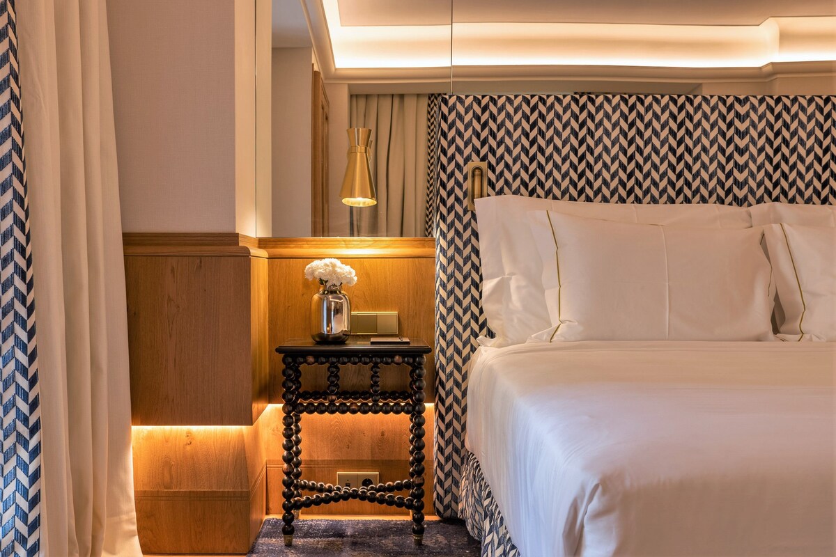 A hotel room with a bed, a nightstand with a lamp, and a patterned wall.