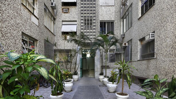 A narrow courtyard with potted plants and a tiled floor, surrounded by a building with patterned walls and multiple windows. - Rio Rentals 021 - C005 Studio em Copa (Rio de Janeiro)