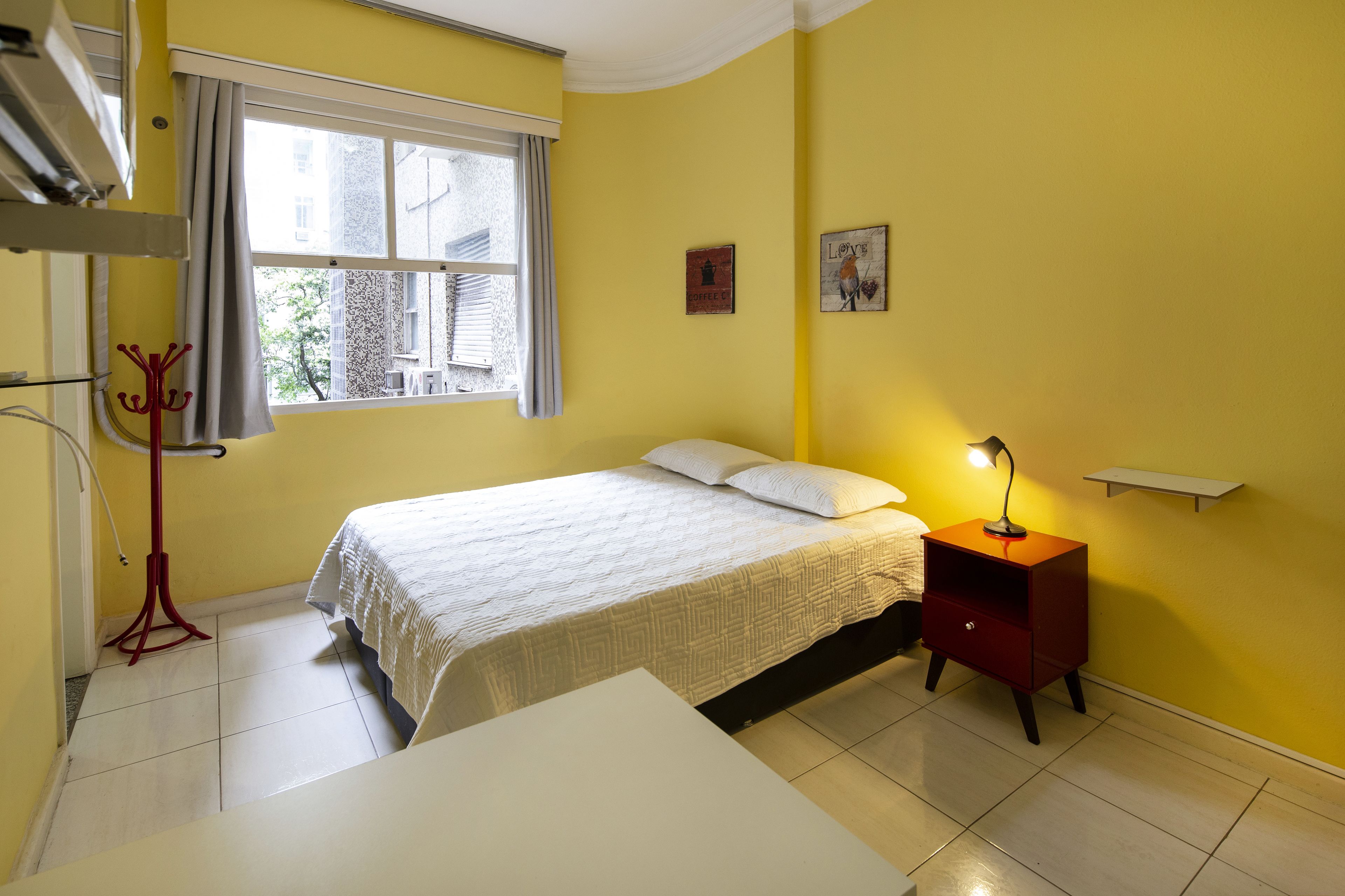 A room with a bed, a bedside table with a lamp, a window with curtains, and a coat rack. - Rio Rentals 021 - C005 Studio em Copa (Rio de Janeiro)