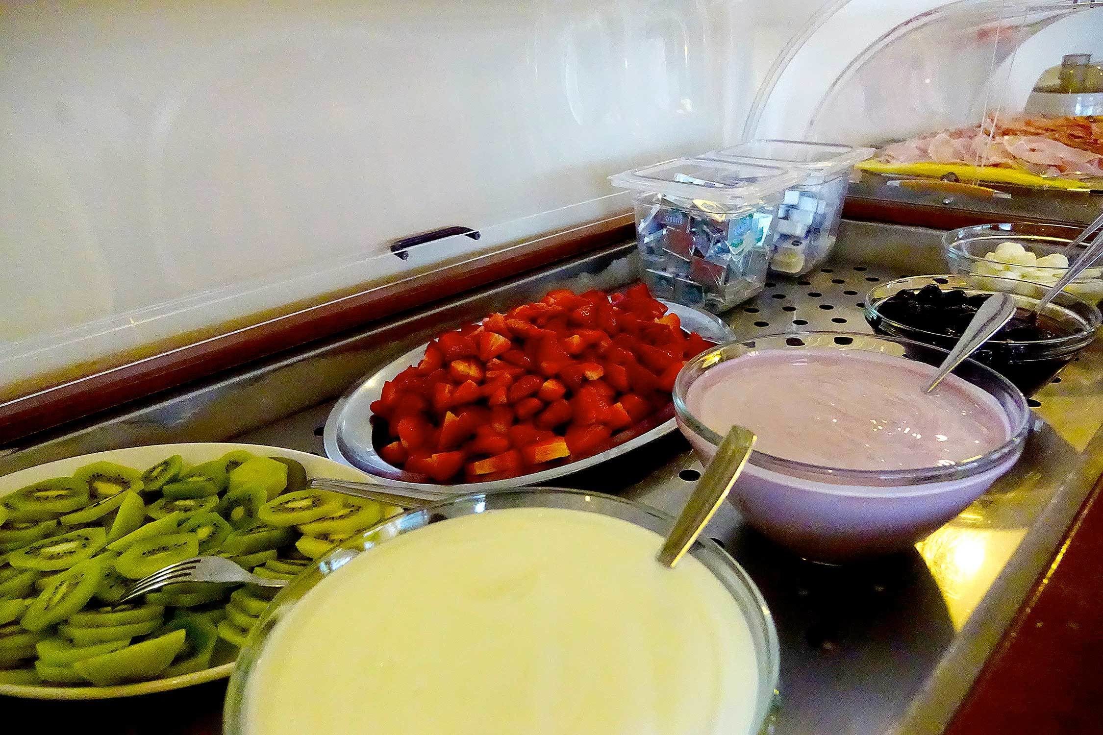 A buffet with various food items including sliced fruits, a bowl of yogurt, and containers of sauces.