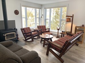 Ein gemütliches Wohnzimmer mit Kamin, Holzmöbeln und großen Fenstern mit Blick auf Bäume.