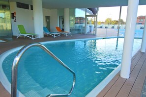 An indoor swimming pool with clear blue water, surrounded by lounge chairs and a wooden deck. - Apartment Alexander Boulevard (Punta del Este)