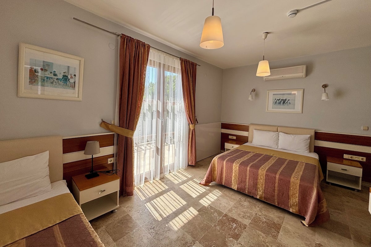 A hotel room with two beds, a large window with curtains, a ceiling fan, and artwork on the walls.