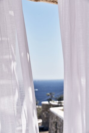 A view through sheer white curtains, revealing a coastal landscape with a clear sky and distant landforms.