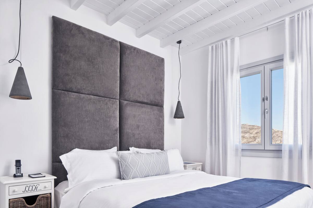 Een moderne slaapkamer met een groot bed, een nachtkastje met een telefoon en een boek, en een raam met gordijnen.