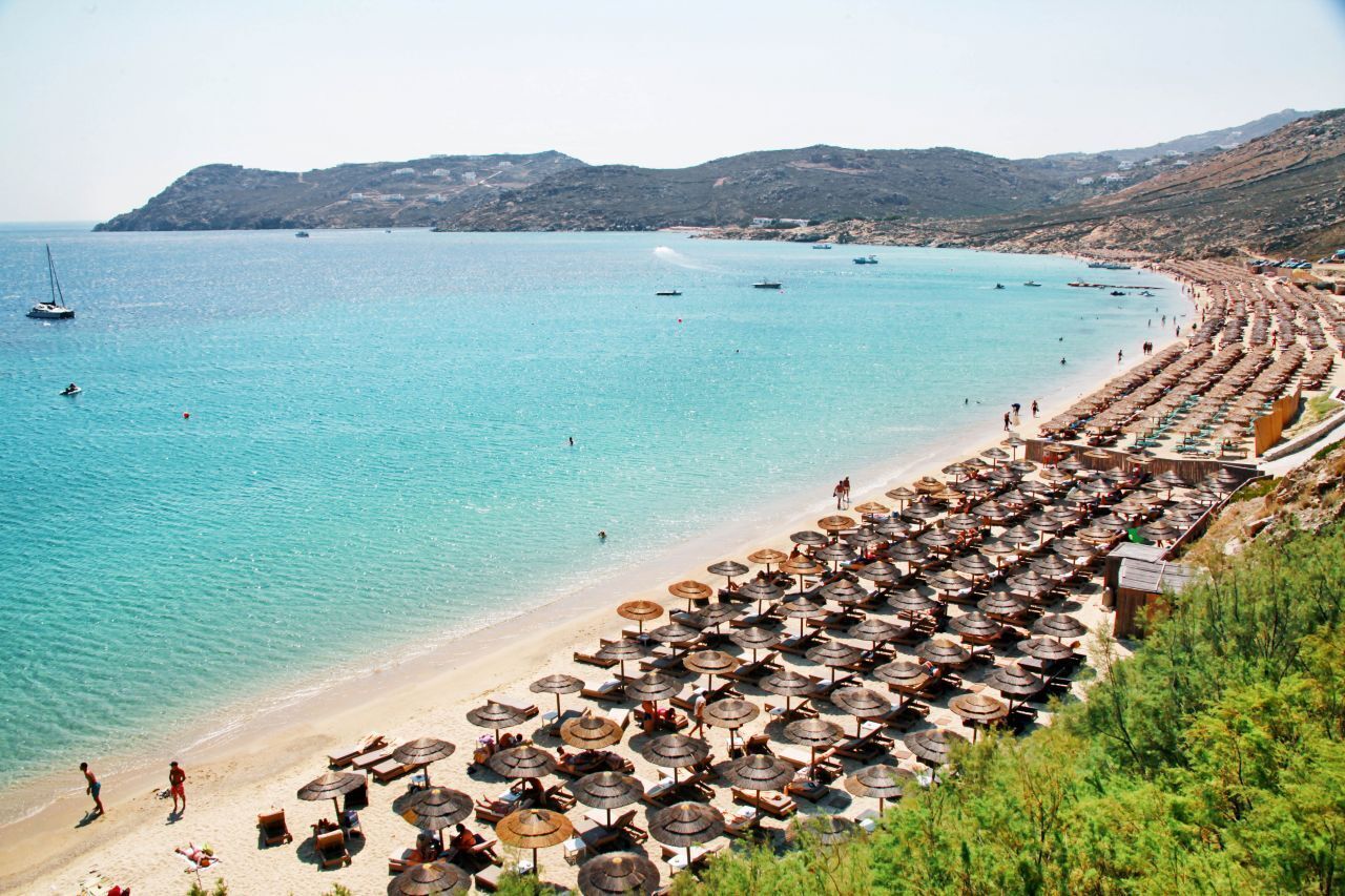 A beach with rows of umbrellas and sun loungers, a clear blue sea, and boats in the distance.