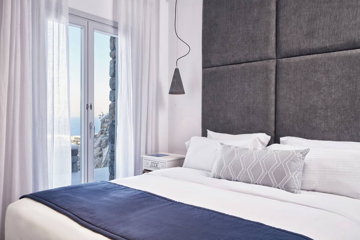 Een moderne hotelkamer met een groot bed, een grijs gestoffeerd hoofdeinde, wit beddengoed en uitzicht door een glazen deur.