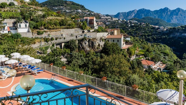 Zona de piscina con sillones de playa y sombrillas, con vistas a una colina con edificios y montañas.