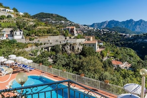 Zona de piscina con sillones de playa y sombrillas, con vistas a una colina con edificios y montañas.