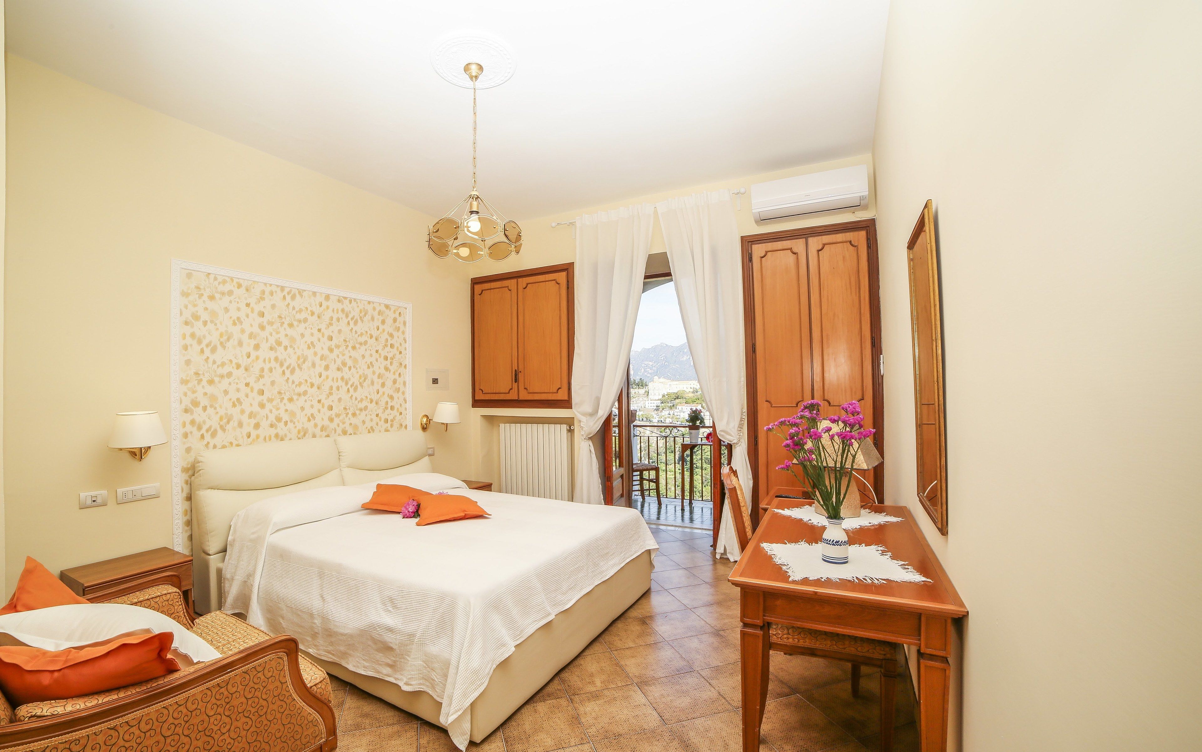 A hotel room with a bed, a chair, a table with a vase, a ceiling light, and a view of the outside.