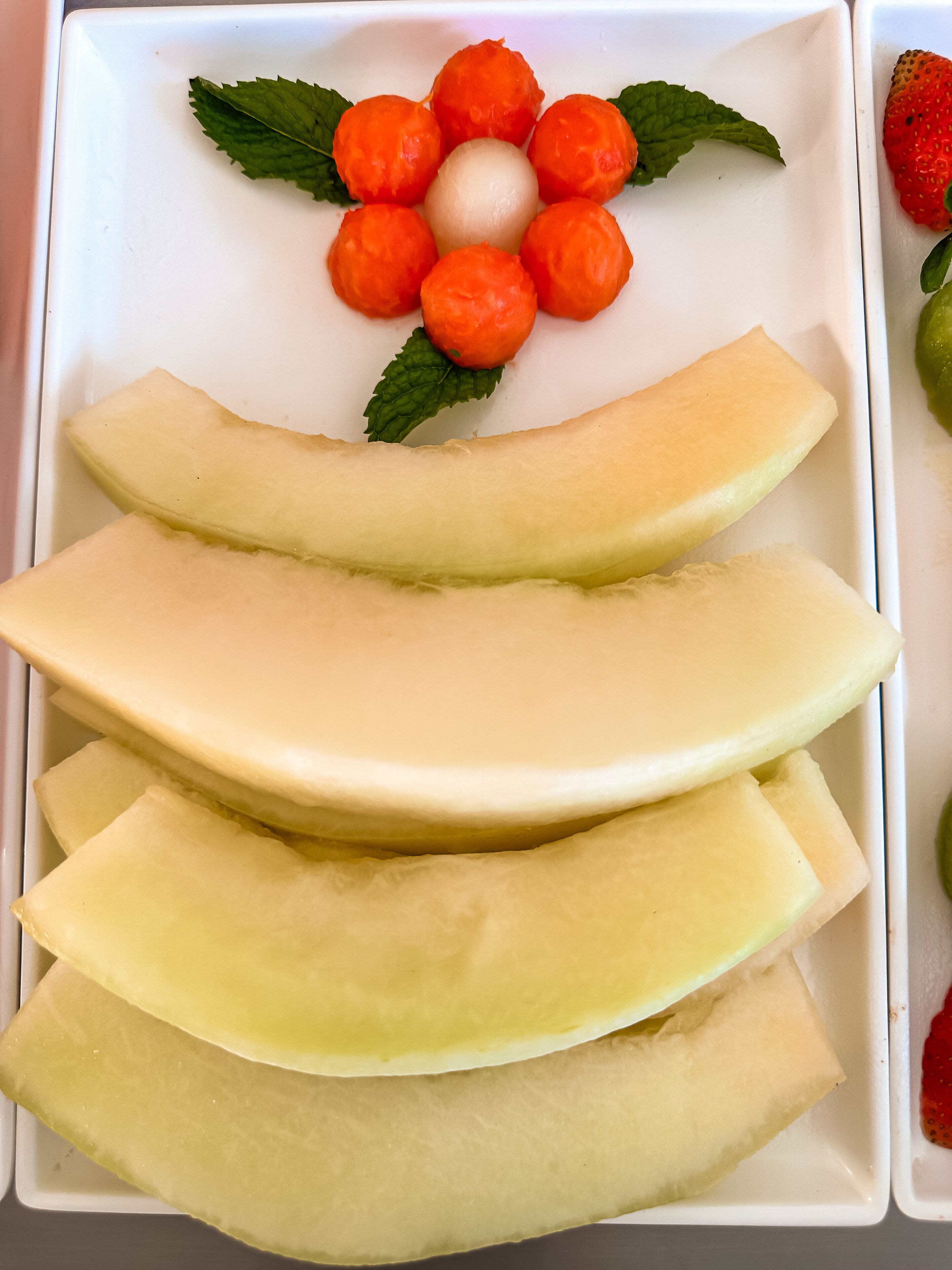 Un piatto bianco rettangolare con melone cantalupo a fette disposto a ventaglio, guarnito con foglie di menta e una fragola.