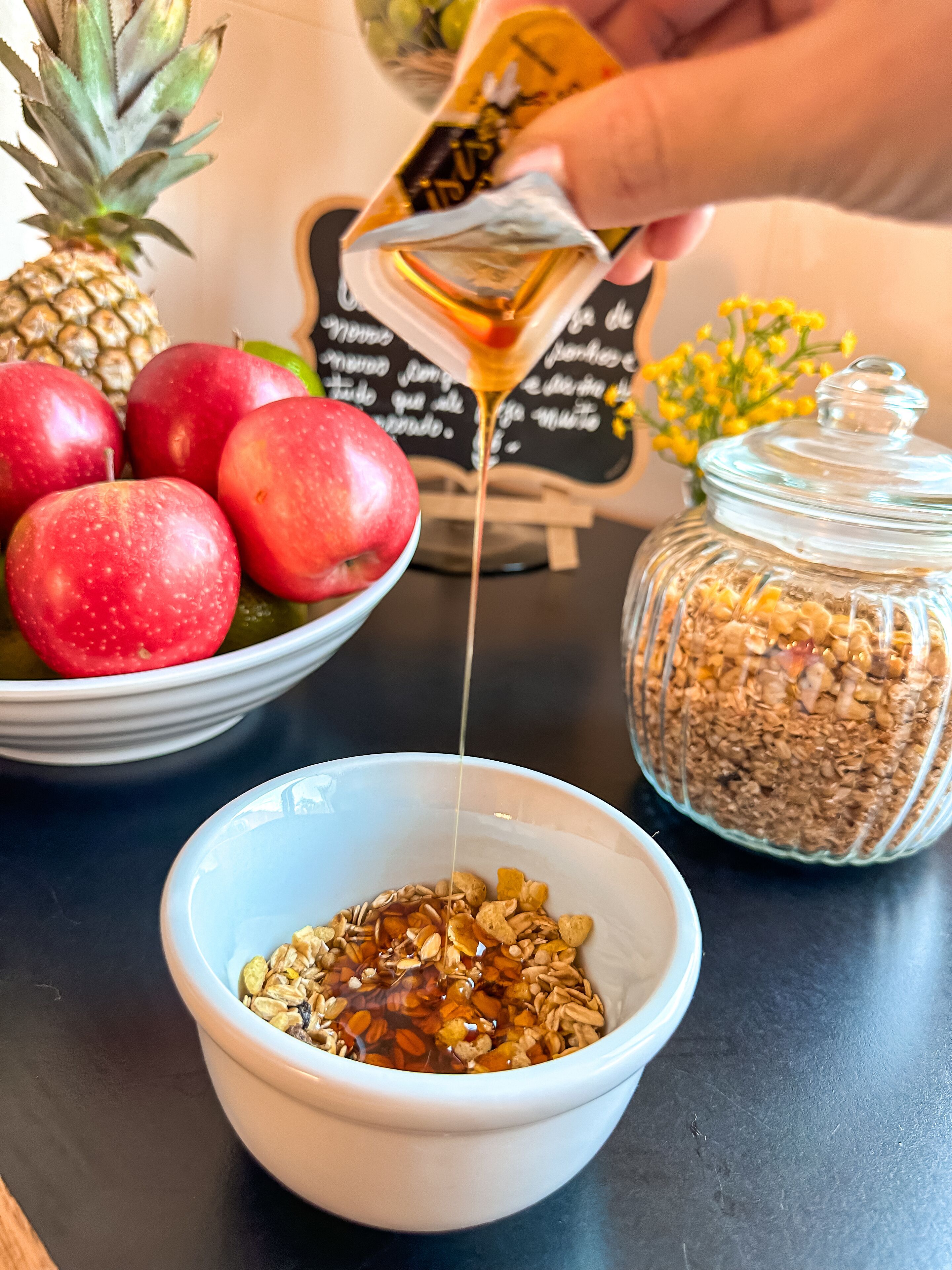 Un tazón de granola con miel siendo vertida, un tarro de granola y un tazón de manzanas rojas.