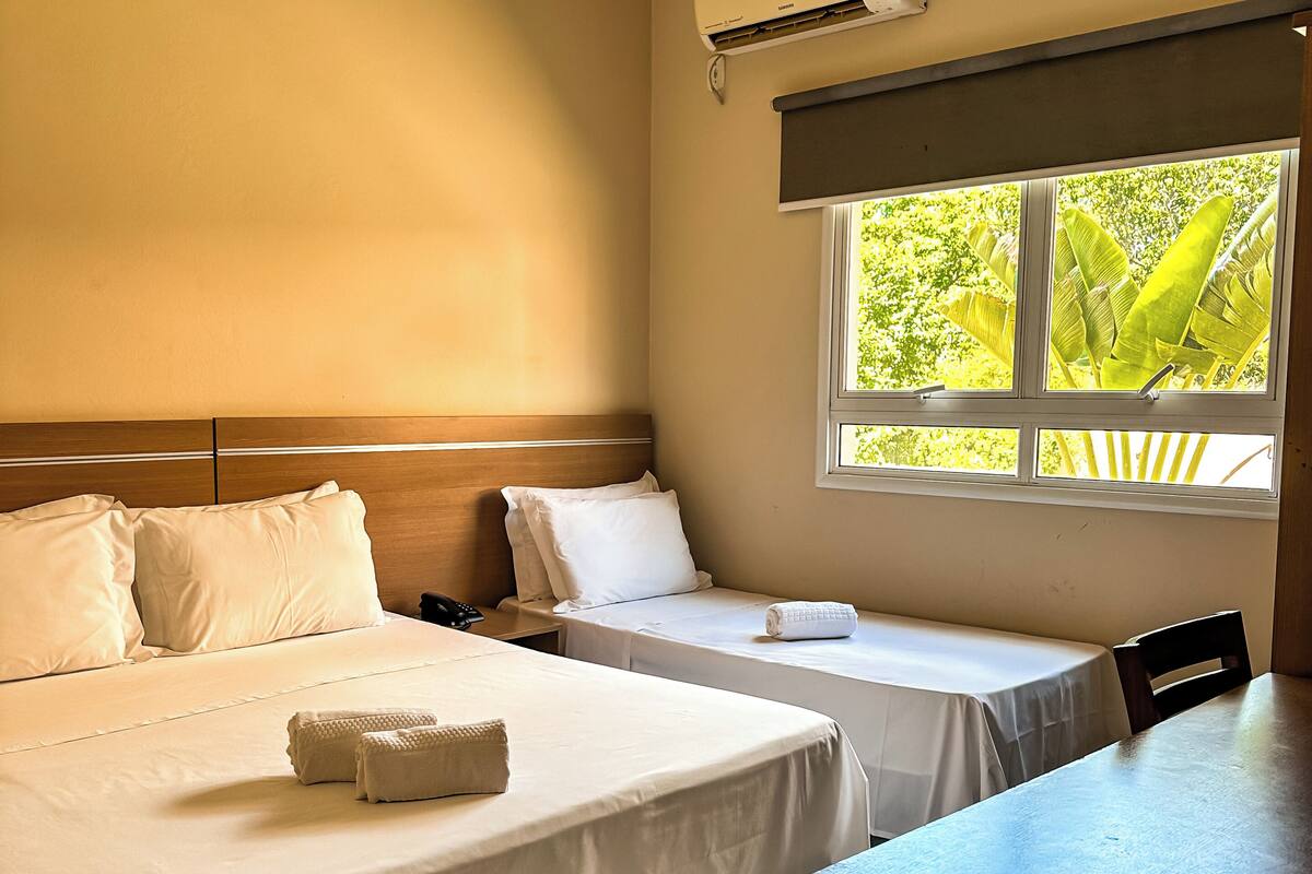 A hotel room with two beds, a desk, and a window with a view of greenery.