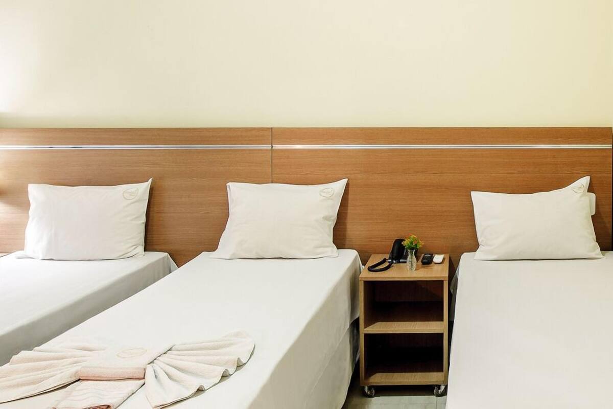 A hotel room with two single beds, a wooden headboard, a nightstand with a phone and a vase of flowers, and a small cart with towels.
