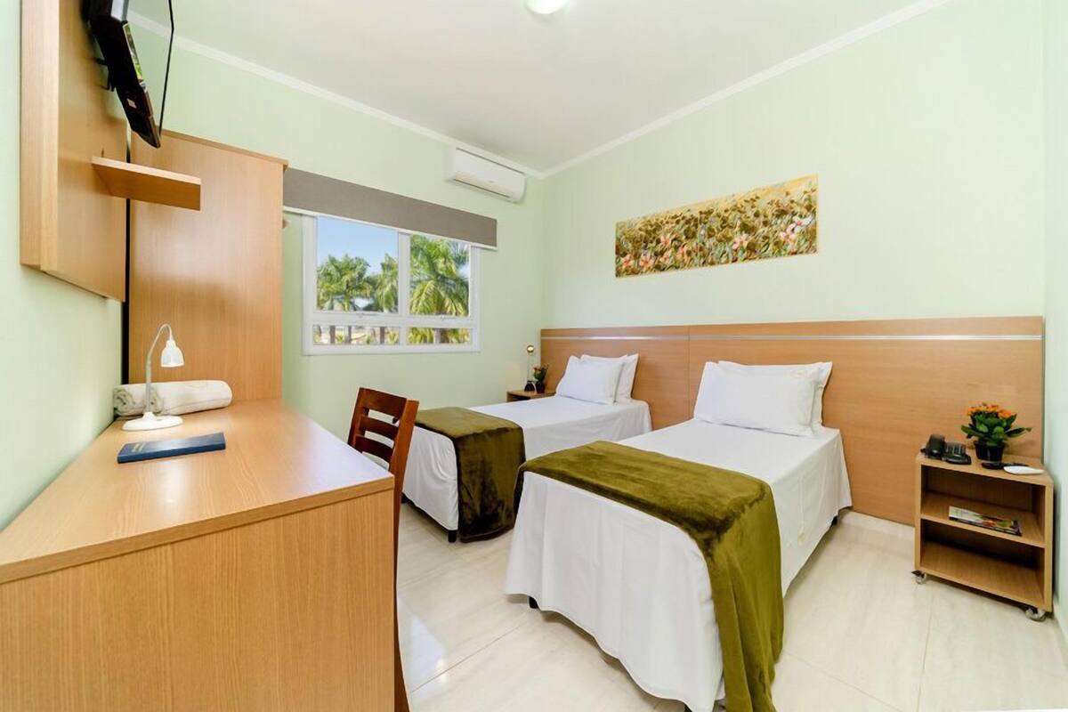 A hotel room with two beds, a desk, a chair, a TV, and a window with a view of palm trees.