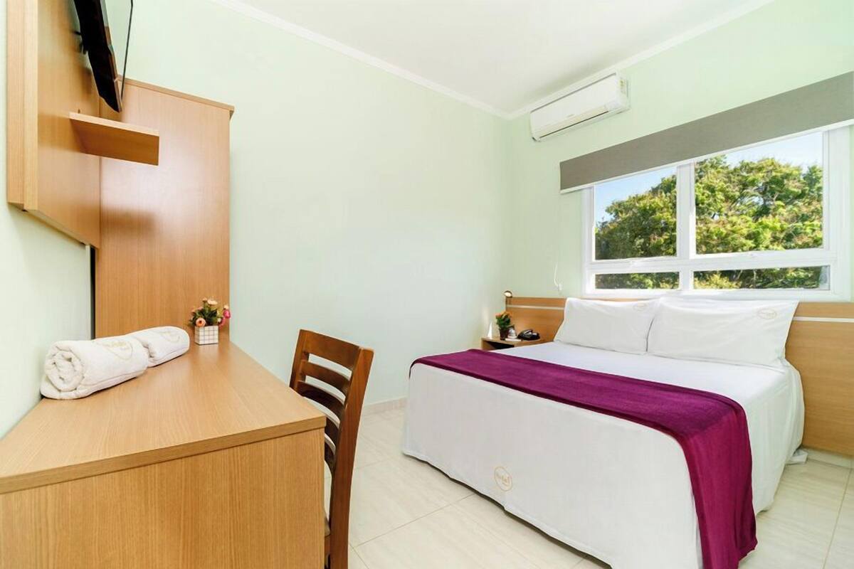 A hotel room with a bed, a desk, a chair, a TV mounted on the wall, and a window with a view of trees.