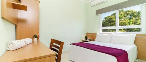 Habitación de hotel con cama, escritorio, silla, televisor montado en la pared y ventana con vista a árboles.