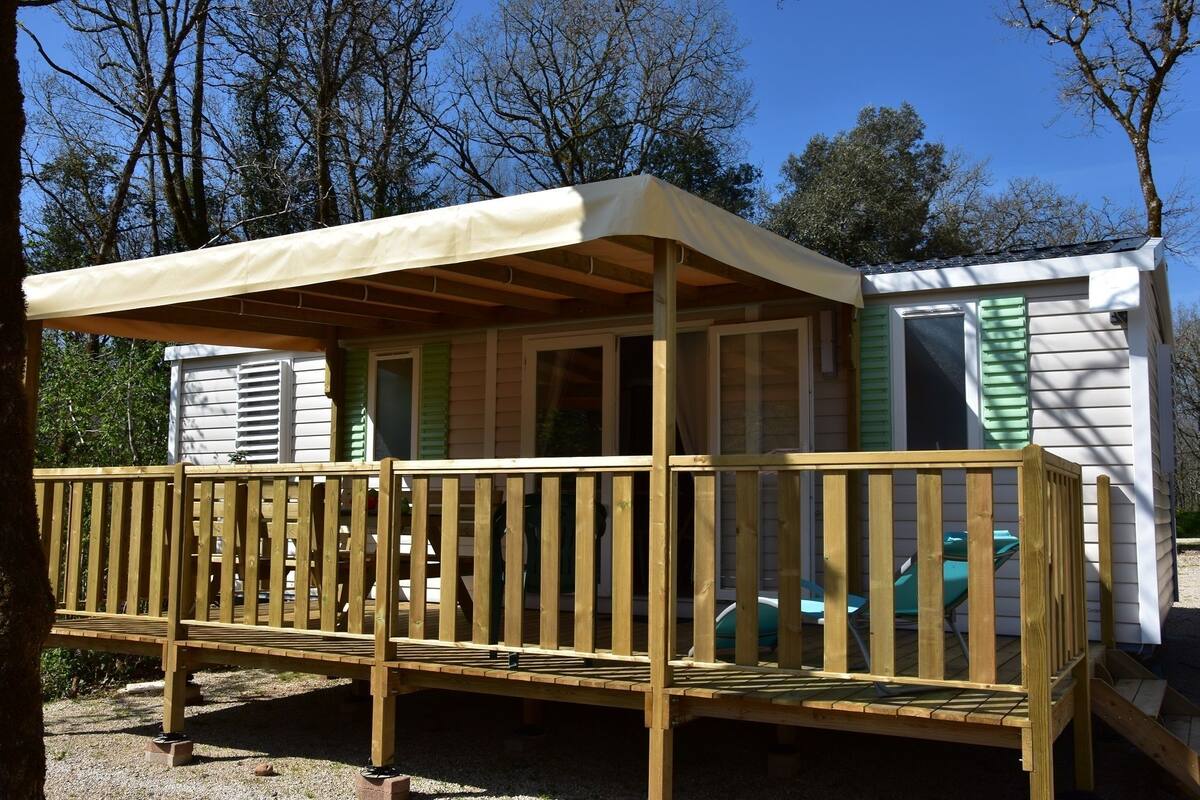 Une terrasse en bois surmontée d’un auvent, un petit bâtiment aux volets verts et un véhicule visible au loin.