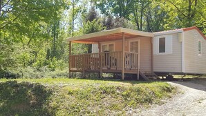 Une maison mobile dotée d’une véranda et d’une petite terrasse, entourée d’arbres et d’herbe.