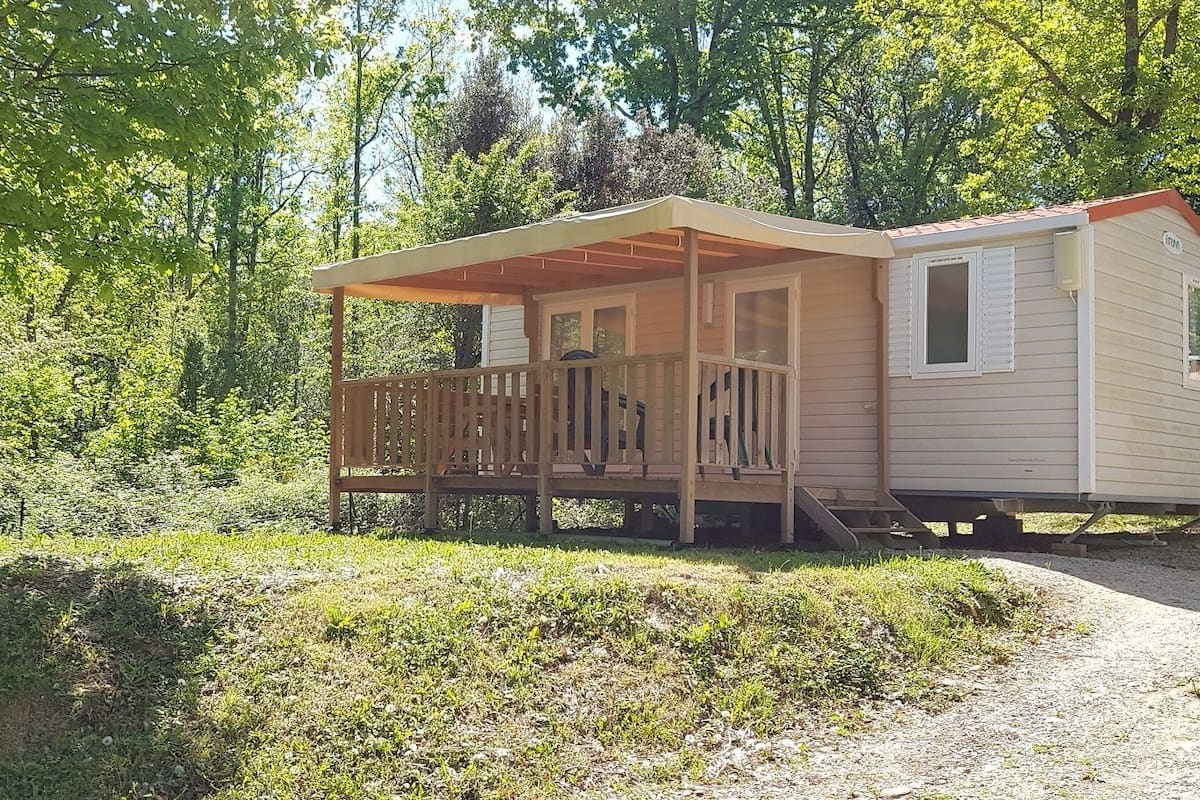 Une maison mobile dotée d’une véranda et d’une petite terrasse, entourée d’arbres et d’herbe.
