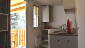 Une petite cuisine avec une fenêtre, un radiateur et un petit plan de travail sur lequel se trouvent une bouilloire et une bouteille rouge.