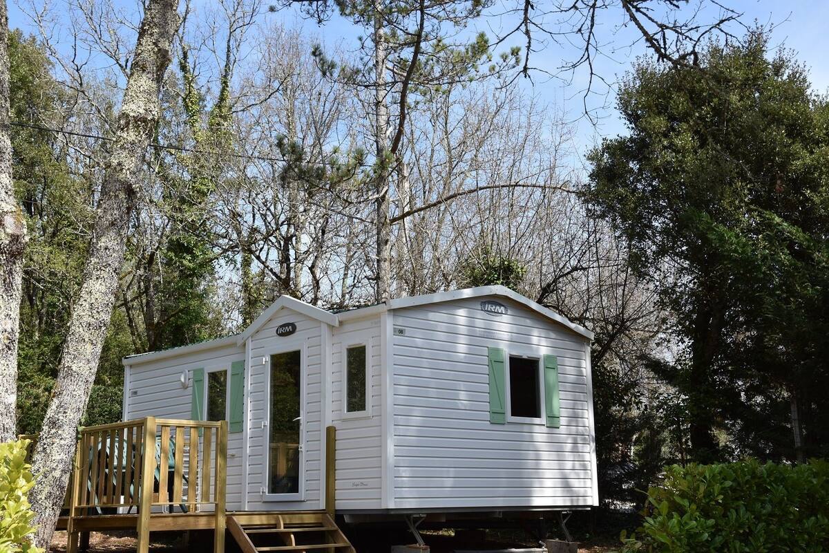Une petite maison mobile blanche, avec des volets verts, une terrasse en bois et une rampe menant à l’entrée.