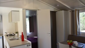 Une cuisine compacte équipée d’une cuisinière à gaz, d’un évier et d’un petit réfrigérateur. On aperçoit un coin repas avec une table et des chaises, ainsi qu’une fenêtre avec des rideaux.