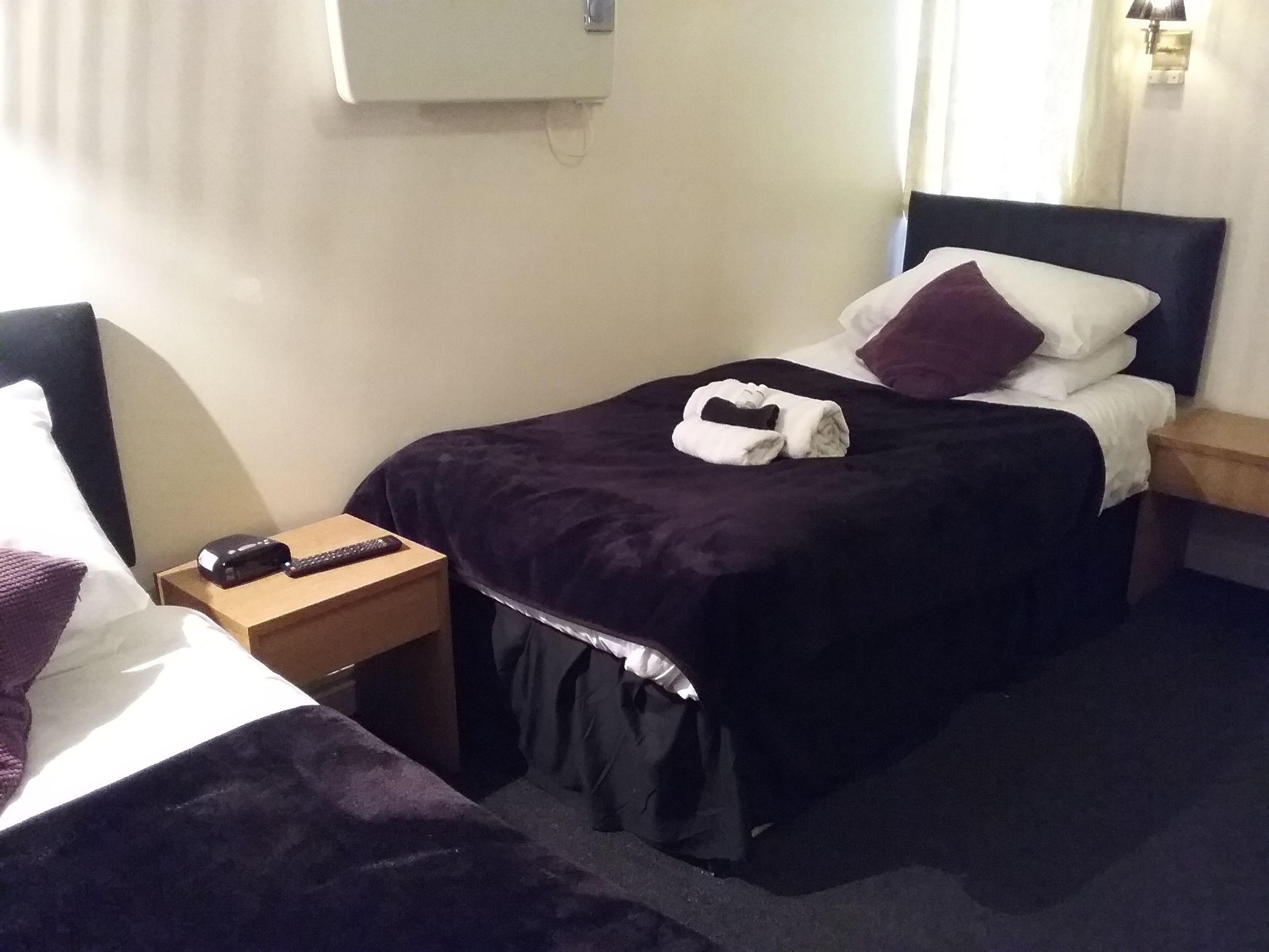 A hotel room with a bed, a nightstand, a remote control, and a wall-mounted air conditioner.
