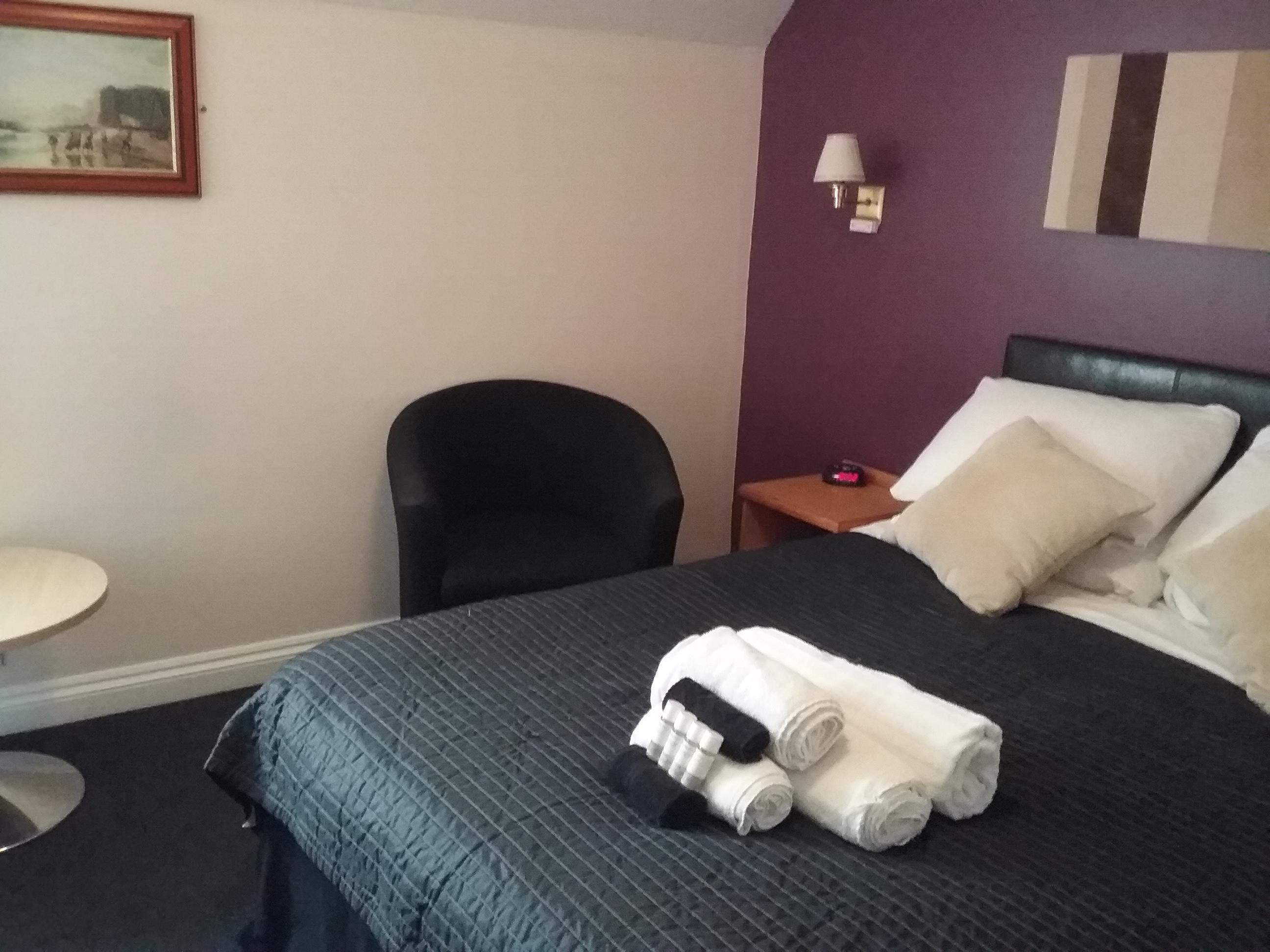 A hotel room with a bed, a chair, a small table, and a painting on the wall.