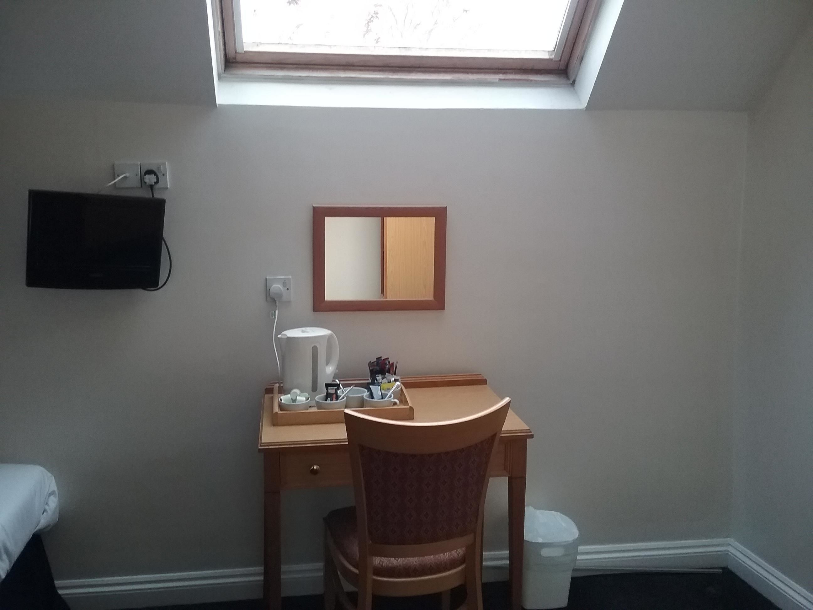 Ein kleines Zimmer mit Schreibtisch, Stuhl und Fenster. An der Wand befindet sich ein Spiegel, auf dem Schreibtisch ein Wasserkocher und eine Tasse. Eine Mülltonne ist in der Ecke sichtbar.
