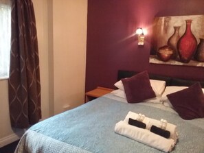 A hotel room with a bed, pillows, a nightstand, a painting, and a window with curtains. - The Prince Consort (Southampton)
