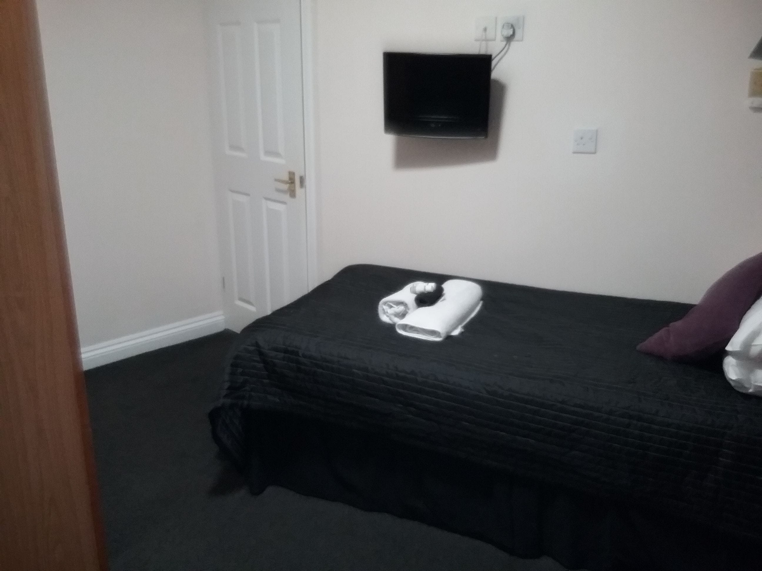 A small, neatly made bed with a black bedspread and a white pillow. A black TV is mounted on the wall above the bed.