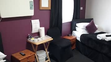 A hotel room with a bed, a desk with a chair, a small table, a trash bin, and a bedside table with a lamp.