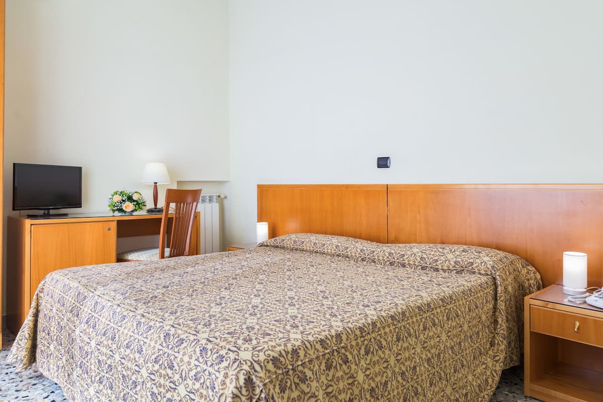 Une chambre d’hôtel avec une tête de lit en bois, un lit recouvert d’une couvre-lit à motifs, un bureau avec une télévision, une lampe et un téléphone.