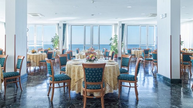 Een eetgedeelte met ronde tafels en stoelen, grote ramen met uitzicht op zee, en een gepolijste marmeren vloer.