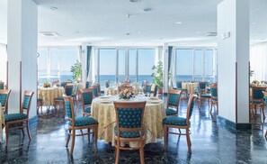 Een eetgedeelte met ronde tafels en stoelen, grote ramen met uitzicht op zee, en een gepolijste marmeren vloer.