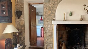 A cozy room with a stone wall, a fireplace, a wooden door leading to a kitchen, and a vintage radio on a dresser.