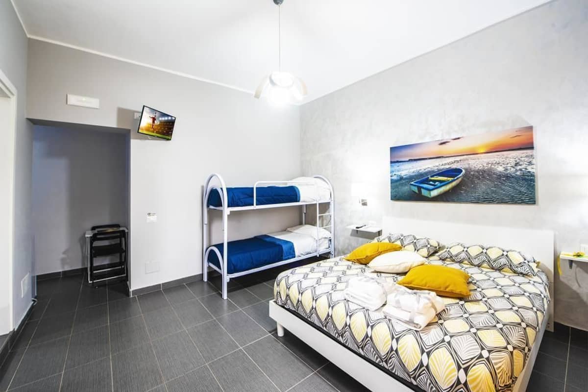 Quarto com beliches, uma cama com roupa de cama estampada, um quadro de um pôr do sol sobre a água e uma mesinha com abajur.