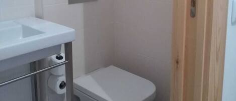 A small, clean bathroom with a white sink, toilet, and mirror. There is a wooden door and a light switch on the wall.