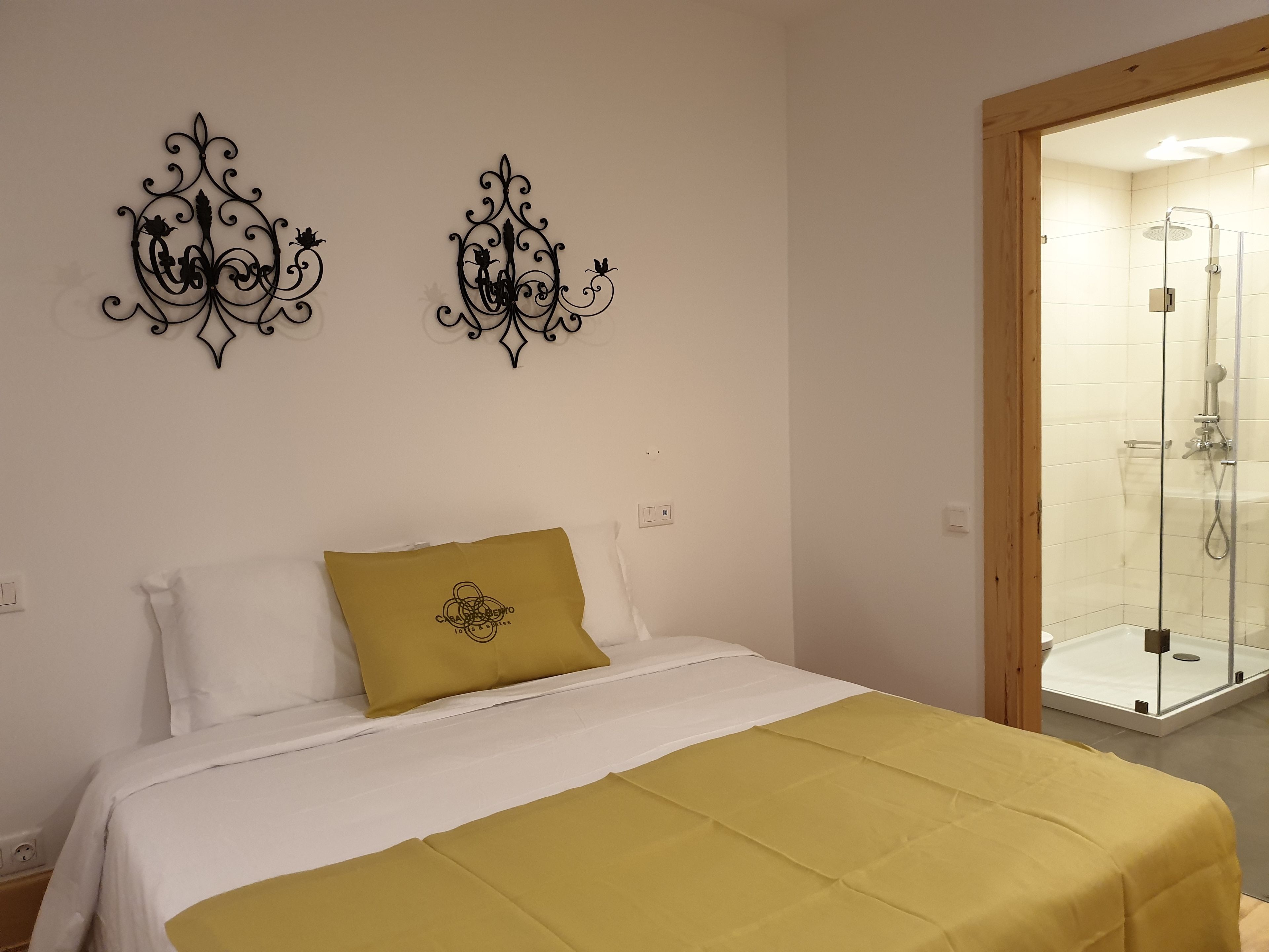 A bedroom with a bed, a yellow throw blanket, two decorative wall pieces, and a glass-enclosed shower area.