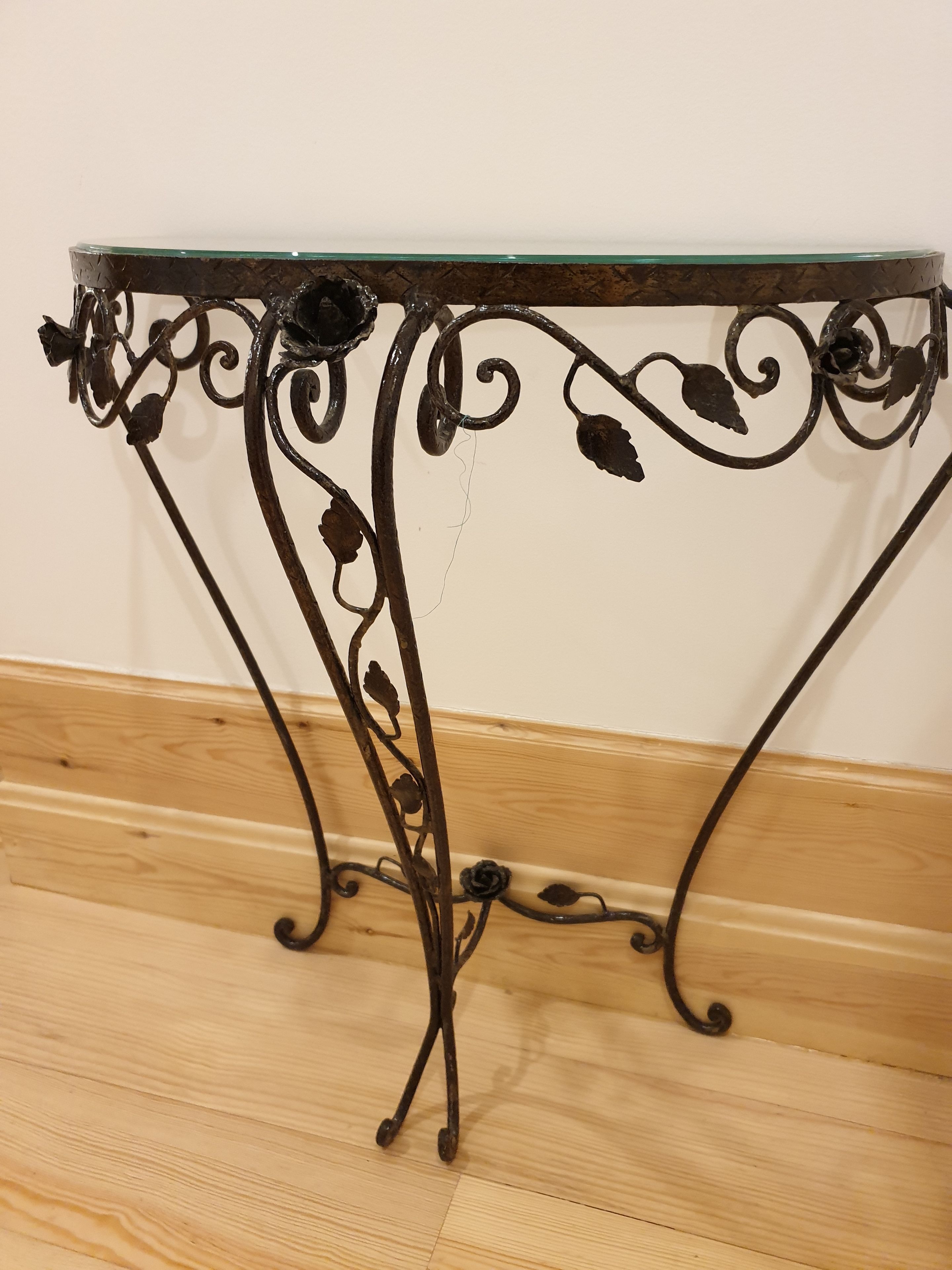 Una mesa metálica con superficie de vidrio y patas ornamentadas frente a una pared lisa.