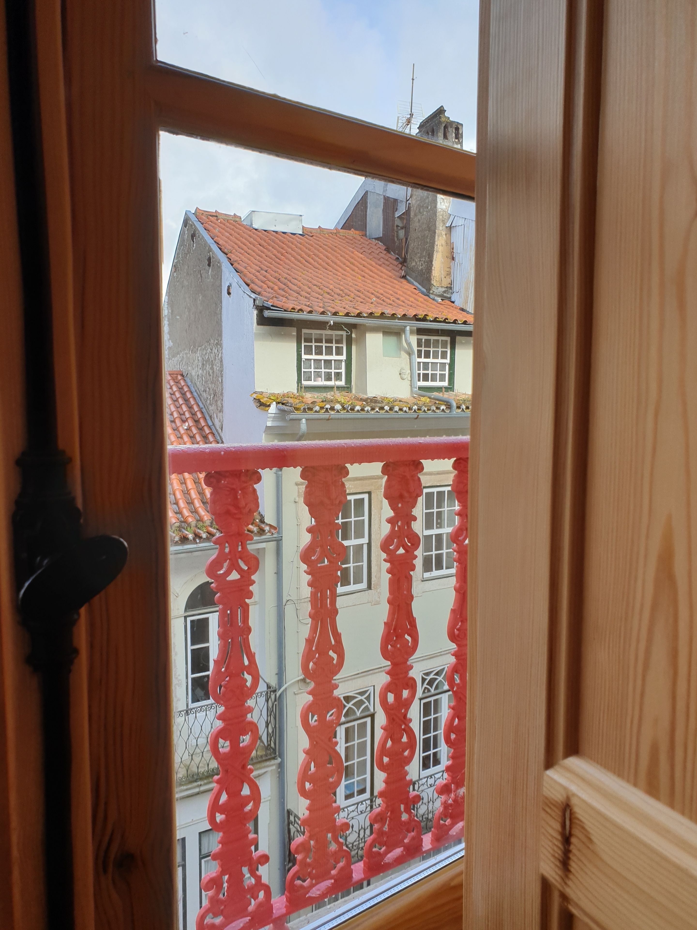 A view through a window with a wooden frame, featuring a balcony with red railings and a building with a red-tiled roof in the background.
