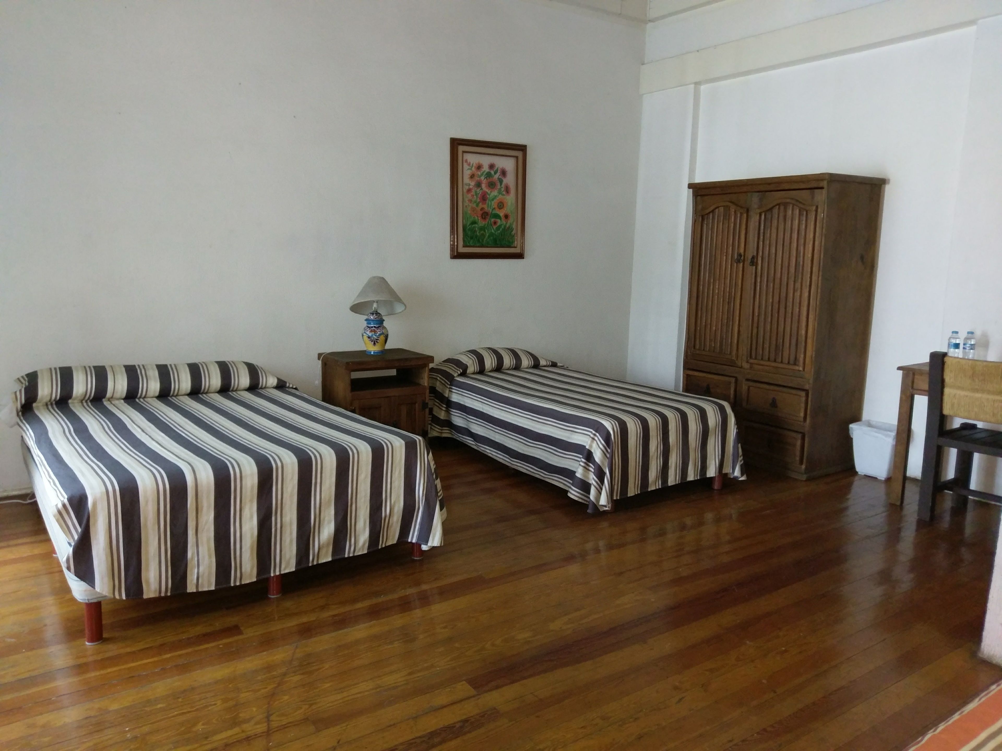 Una habitación con dos camas, un armario de madera, una mesita con una lámpara y un cuadro en la pared.