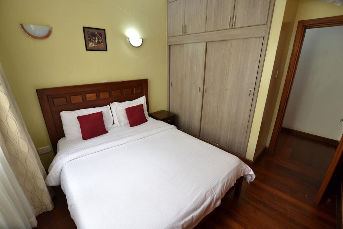 A bedroom with a wooden headboard, a bed with white bedding and red pillows, a wardrobe, and a nightstand with a lamp.