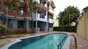 A swimming pool with a metal ladder, surrounded by a brick building with balconies and palm trees. - Lighthomez Angles Heights (Nairobi)