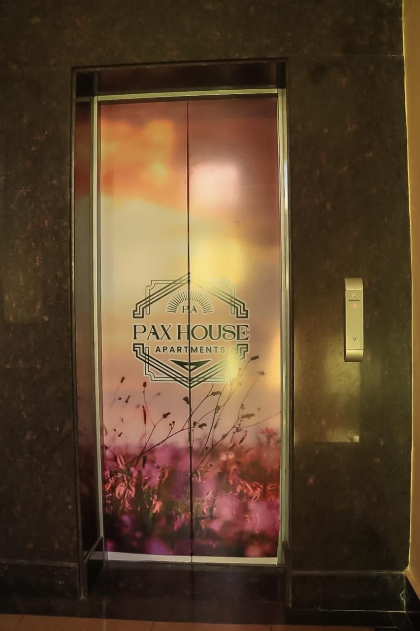 An elevator with a glass door featuring the text 'PAX HOUSE APARTMENTS' and a decorative floral design.