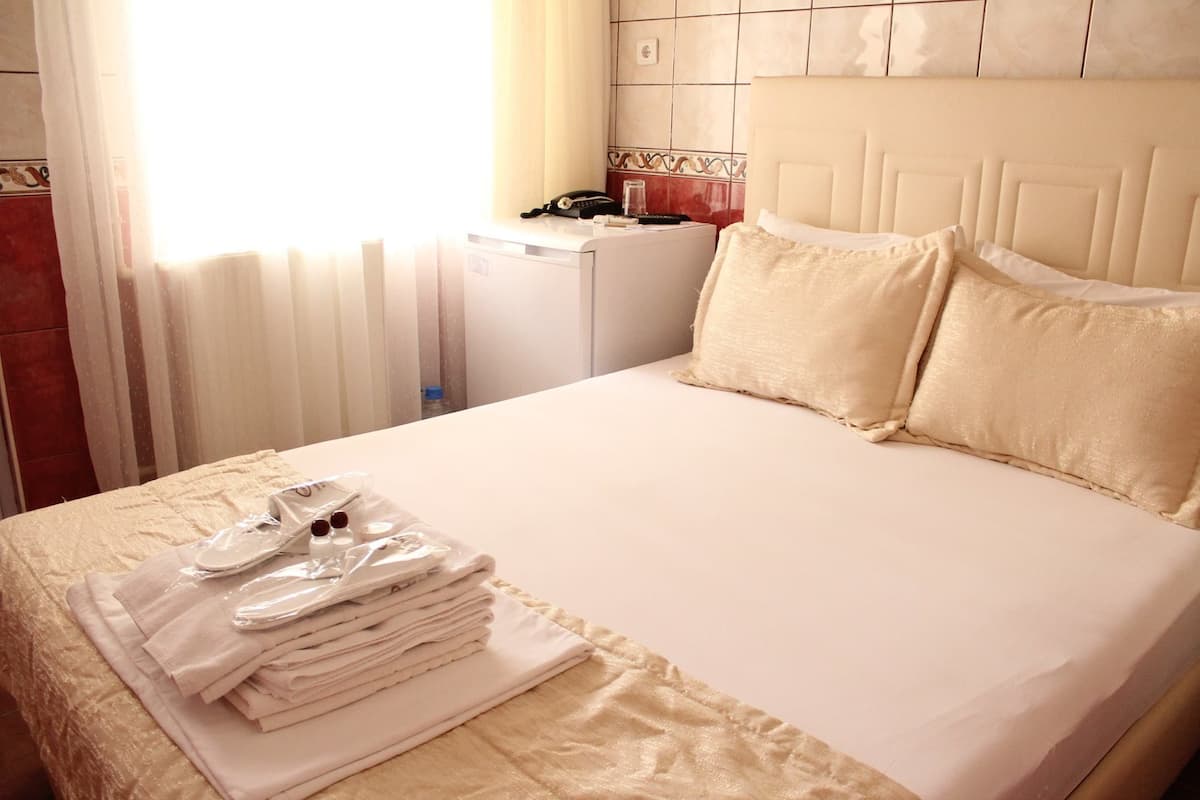 Een net opgemaakt bed met een witte deken en kussens, een klein nachtkastje met een telefoon en een glas water, en een stapel opgevouwen handdoeken op het bed.