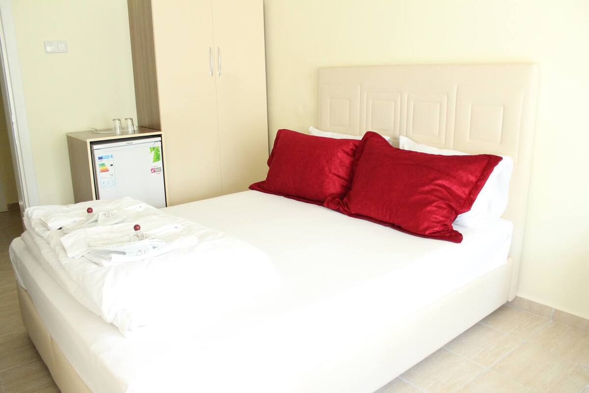 Een netjes opgemaakt bed met witte lakens en twee rode kussens, een klein nachtkastje met een lamp, en een roomkleurige kast in een hotelkamer.