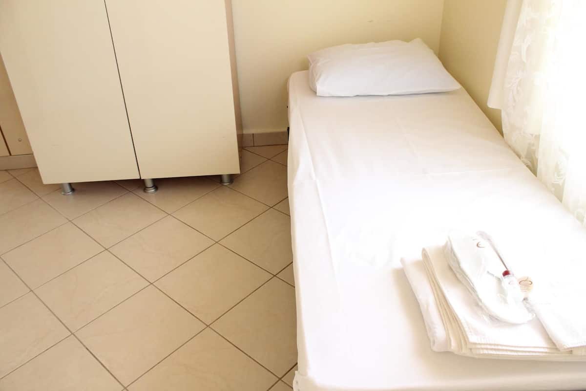Een enkel bed met een witte matras en kussen, een klein nachtkastje met een opgevouwen witte handdoek en een wit voorwerp, en een gesloten witte kast.